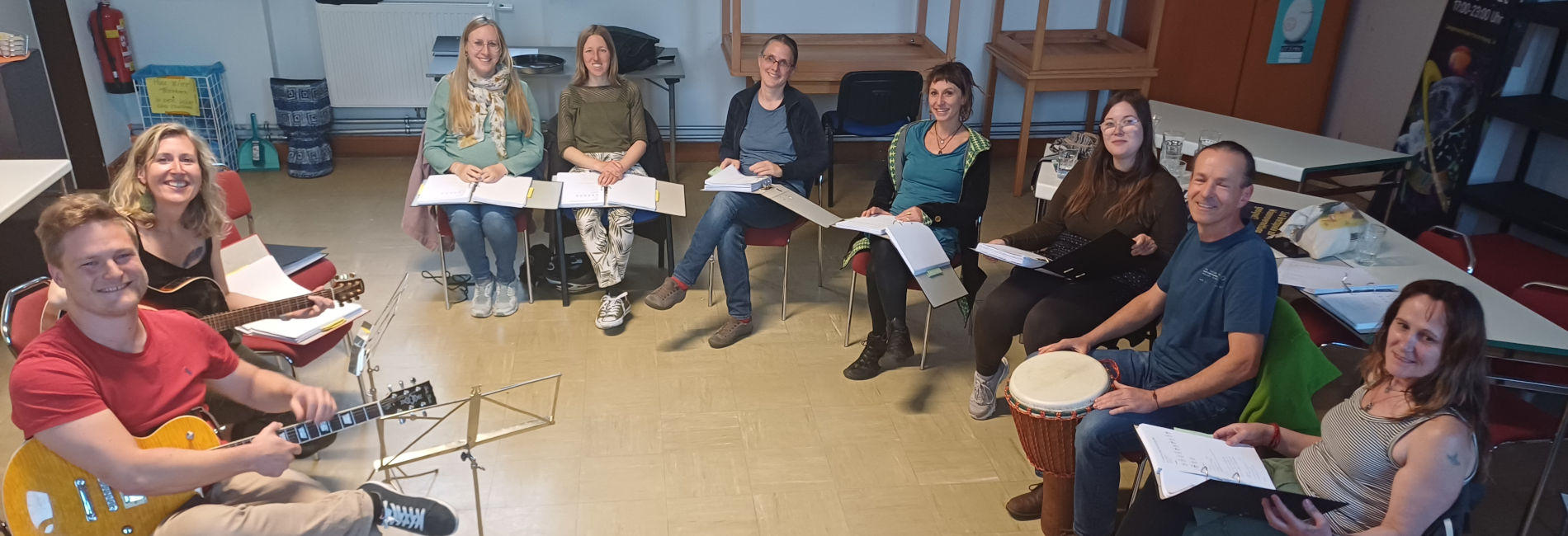 Eine Runde von Menschen mit Gitarren oder Songtexten in der Hand im Sesselkreis lächtel in die Kamera
